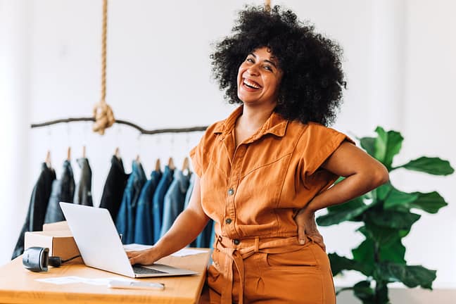 Black small business owner smiling at the camera while standing in her shop. Happy businesswoman managing her clothing orders on a laptop. Young female entrepreneur running an online clothing store.